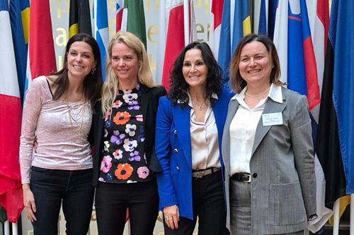 Four people standing in front of a display of multiple national flags in a formal setting.