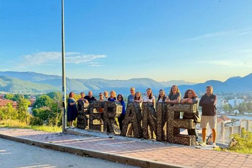 A group of about 15 people posing behind large decorative letters spelling \"BERANE\" on a scenic lookout, with mountains and the city in the background.
