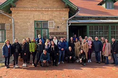 A group of people standing in front of a brick building with green‑framed windows and a sign reading ‘EKO CENTAR’.