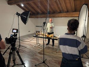 A heritage holder presents traditional food products arranged on a table while participants record the demonstration with a camera, studio lights and a reflector during the THRIVE Academy.