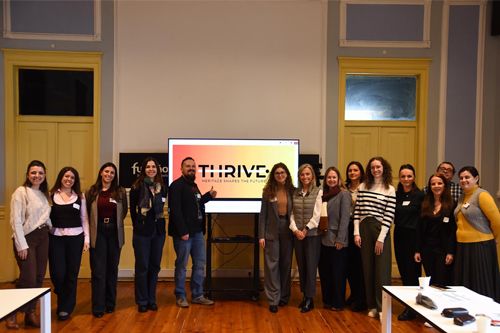 A group of thirteen people standing indoors in front of a screen displaying the title ‘THRIVE \'.