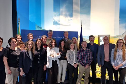 A group of people standing indoors in front of a blue backdrop with European-themed graphics and a flag.