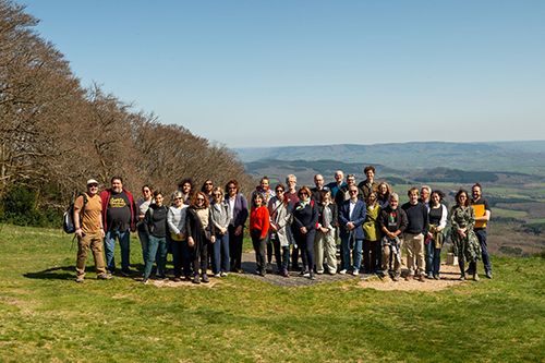 A group of people standing on a grassy area with trees and distant hills under a clear sky.