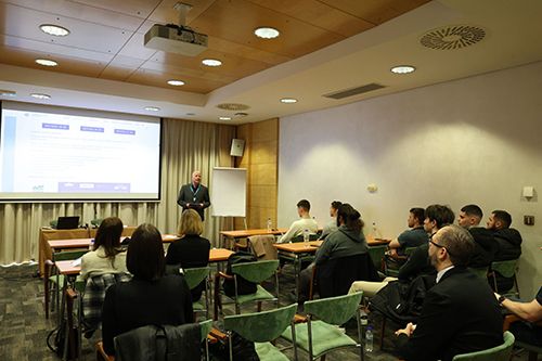 People seated in a classroom-style room facing a presenter standing beside a projection screen.